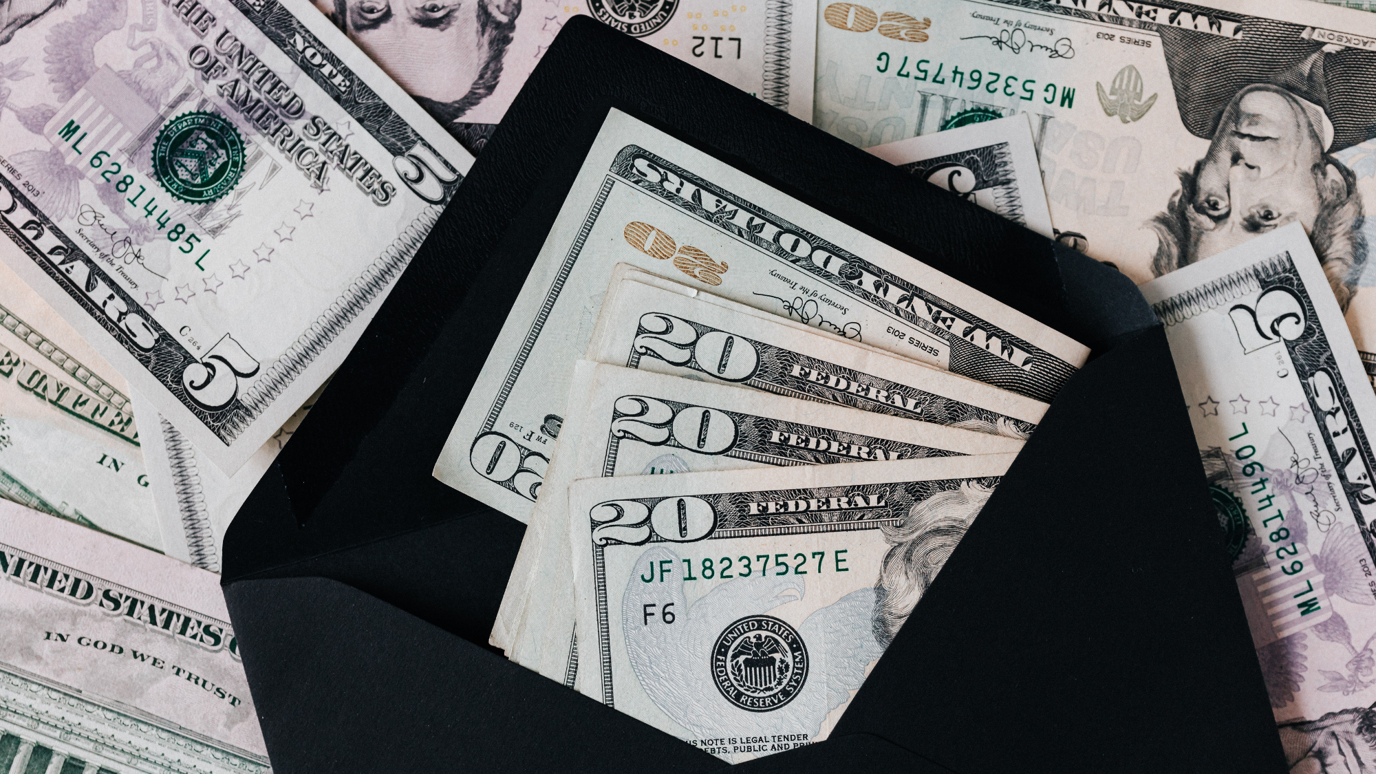 A black envelope filled with twenty dollar bills with many bills in the background.