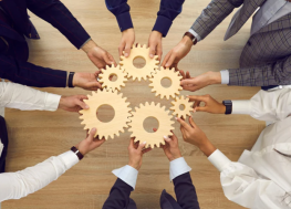 Overhead view of working corporate professionals holding wooden gears together around a table, symbolizing teamwork, collaboration, and shared strategy.