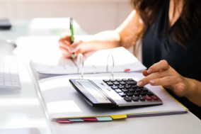 Woman in a dark navy sleeveless shirt uses a calculator while writing in a binder, with tabs, paperwork, and a keyboard on a bright white desk.