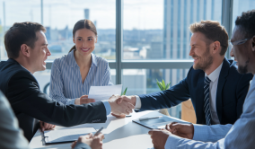 Four business professionals in navy suits shake hands at a bright glass meeting table with white papers and blue city views.