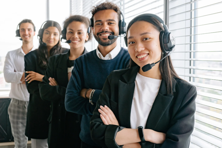 Five smiling co-workers in black blazers and headsets stand by white blinds in a bright office with smooth glass windows.